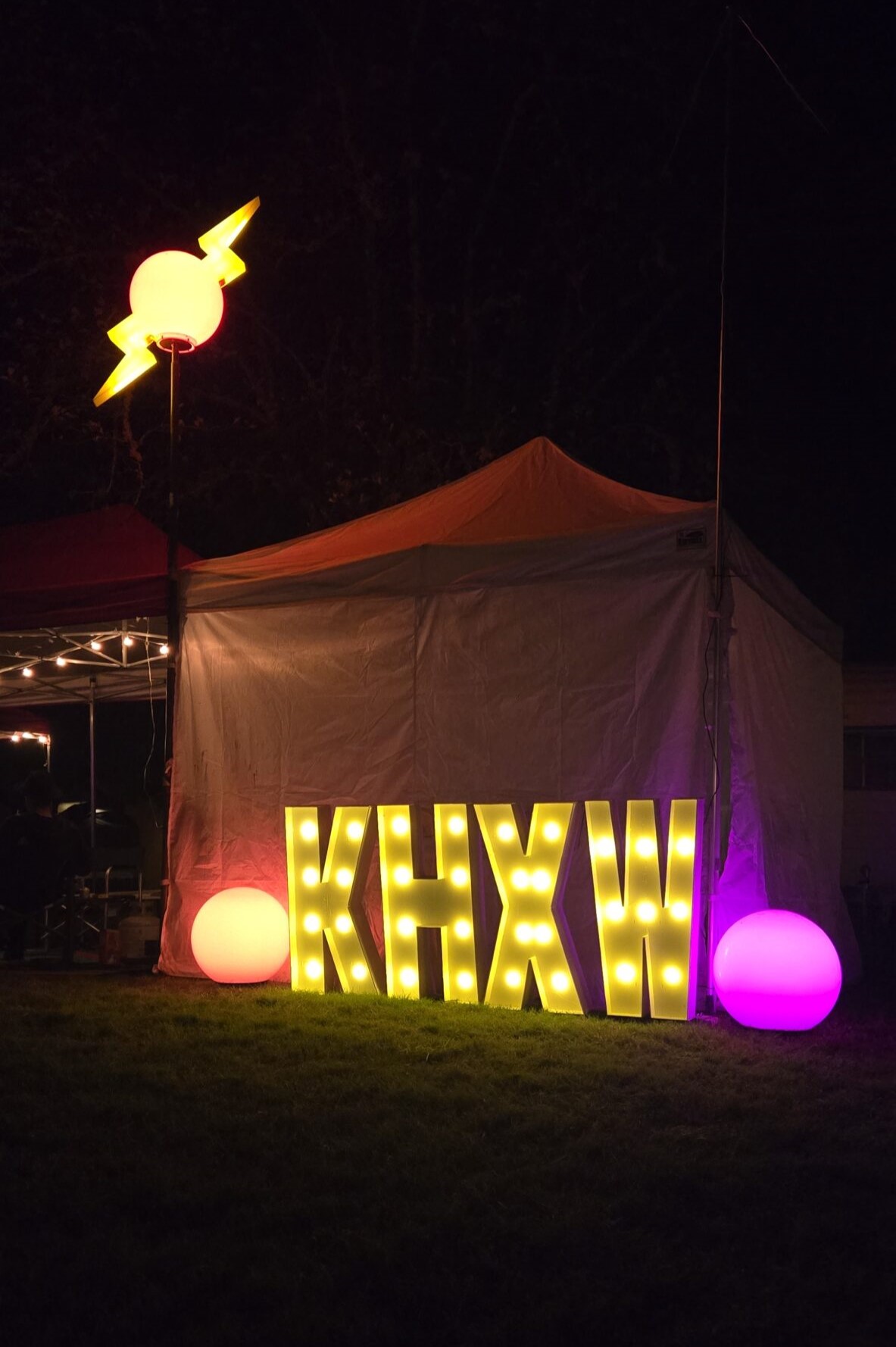 A nighttime view of the letters KHXW lit up in front of a popup shelter. There are colored globes decorating either side of the sign, and a lighted yellow beacon on a pole above the tent that looks like a globe with lightning bolts emerging.